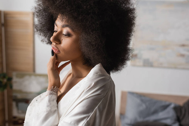 riflessivo donna afro-americana in abito bianco satinato labbro toccante mentre in piedi a casa - Foto, immagini