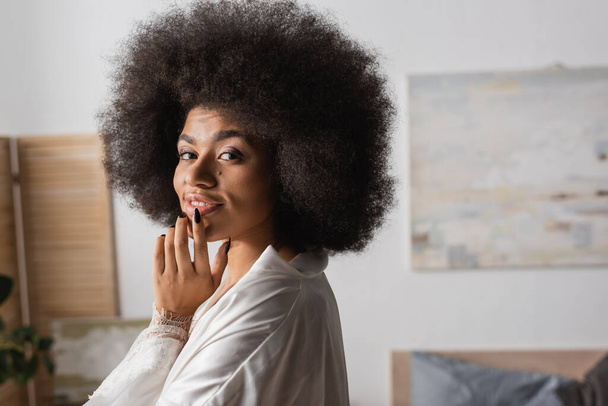 séduisante femme afro-américaine en robe de soie blanche touchant la lèvre et souriant à la caméra à la maison - Photo, image