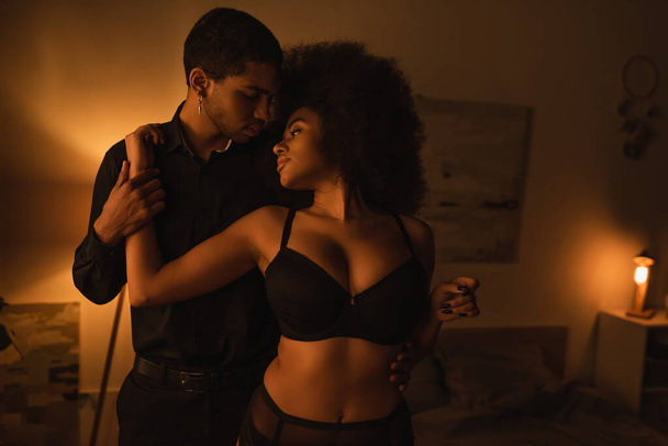 sensual african american woman in black underwear hugging young boyfriend in dark room with luminous lamp - Photo, image