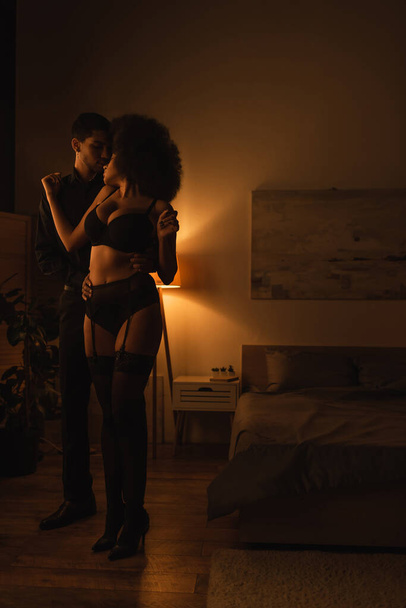 pleine longueur de femme afro-américaine en sous-vêtements noirs debout près du jeune homme dans la pièce sombre avec éclairage - Photo, image