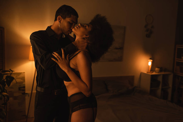 side view of sensual african american woman in sexy lingerie and man in black shirt hugging in dark bedroom  - Foto, Imagen