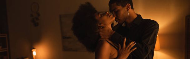 πλαϊνή άποψη του Αφροαμερικανού άνδρα σε μαύρο πουκάμισο αγκαλιάζει παθιασμένη γυναίκα σε σκοτεινό δωμάτιο με φωτισμό, πανό - Φωτογραφία, εικόνα