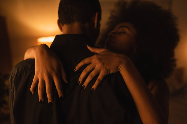 спокуслива афроамериканська жінка обіймає чоловіка в чорній сорочці в темній кімнаті з освітленням
 - Фото, зображення
