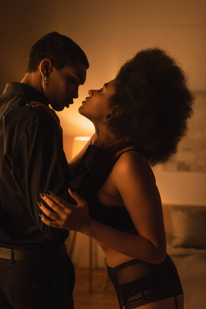 vista lateral de la apasionada mujer afroamericana cerca de hombre en camisa negra en habitación oscura con iluminación - Foto, Imagen