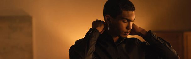 young african american man adjusting collar of black shirt in evening at home, banner - Foto, Imagen