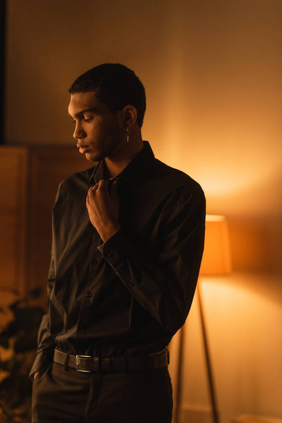 morena hombre afroamericano en camisa negra de pie con la mano en el bolsillo de los pantalones en la habitación con lámpara luminosa - Foto, imagen