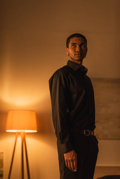 jeune homme afro-américain en vêtements noirs regardant loin tout en se tenant dans une pièce sombre avec une lampe lumineuse - Photo, image