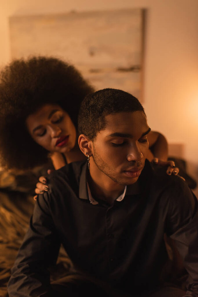 Siyah gömlekli adamın omuzlarına dokunan kıvırcık Afrikalı Amerikalı kadın geceleri yatak odasında oturuyor. - Fotoğraf, Görsel