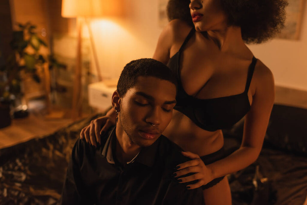african american woman in black bra embracing shoulders of boyfriend sitting in bedroom with closed eyes - Fotoğraf, Görsel