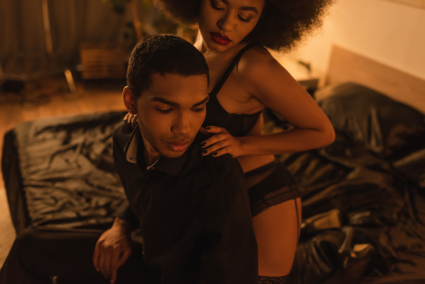 sexy african american woman in underwear hugging shoulders of young man sitting on bed at night - Foto, Imagen