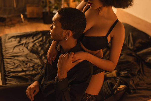 mladý africký Američan muž drží ruku sexy přítelkyně, zatímco sedí na černé satén lůžkoviny - Fotografie, Obrázek