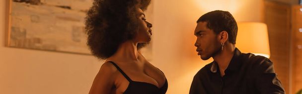 сексуальная африканская американка в лифчике рядом с молодым парнем в черной рубашке в спальне дома, баннер - Фото, изображение