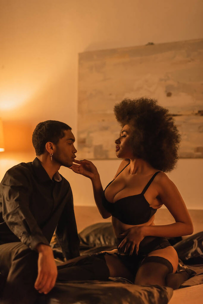 вид сбоку страстной африканской женщины в сексуальном нижнем белье и молодого человека на кровати вечером - Фото, изображение