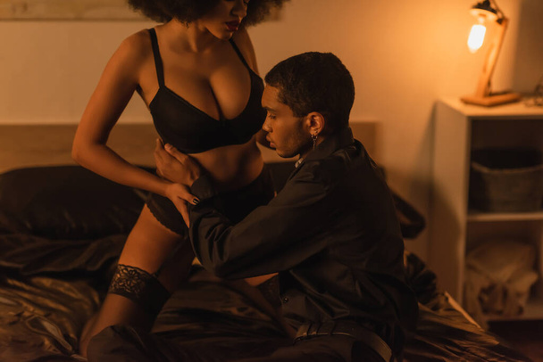 young man embracing passionate african american woman in black underwear in dark bedroom with luminous lamp - Foto, afbeelding