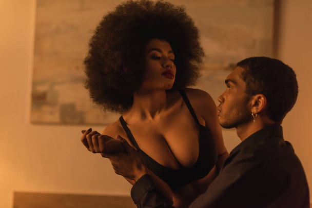 young man holding hand of sexy african american woman in black bra in bedroom - Foto, Imagem
