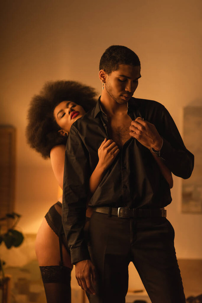 femme afro-américaine passionnée déshabiller homme en chemise noire et pantalon dans la chambre à coucher la nuit - Photo, image