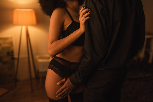 vue recadrée de l'homme en vêtements noirs près sexy afro-américaine petite amie en lingerie noire - Photo, image