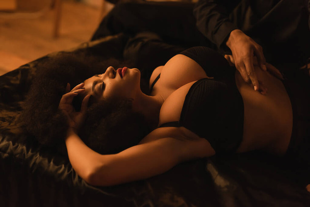 young man touching seductive african american woman lying on silk bedding with closed eyes - Photo, image