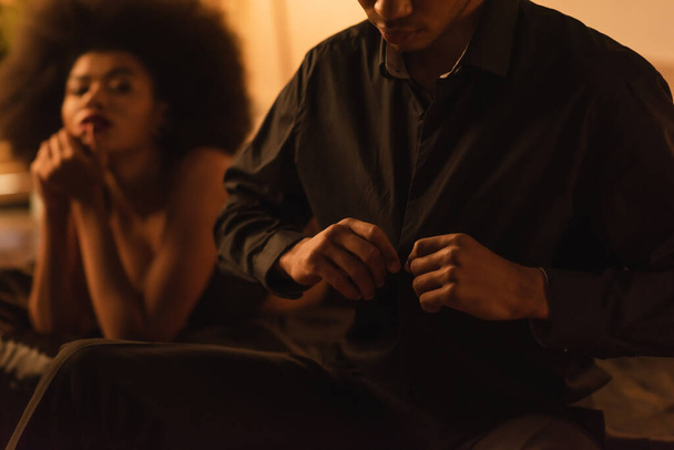 θολή αφρικάνικη αμερικανική γυναίκα κοιτάζοντας τον άνδρα ξεκούμπωμα μαύρο πουκάμισο στην κρεβατοκάμαρα - Φωτογραφία, εικόνα