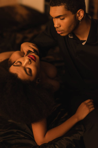 młody afrykański amerykański mężczyzna patrząc na namiętną kobietę leżącą z zamkniętymi oczami na czarny satynowy pościel - Zdjęcie, obraz