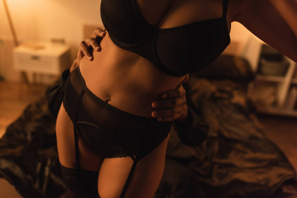 vue recadrée de femme américaine africaine sexy en sous-vêtements noirs près de l'homme l'embrassant dans la chambre sombre - Photo, image