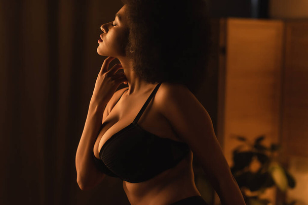 side view of young african american woman with sexy bust touching neck while posing at night in bedroom - Foto, Imagem