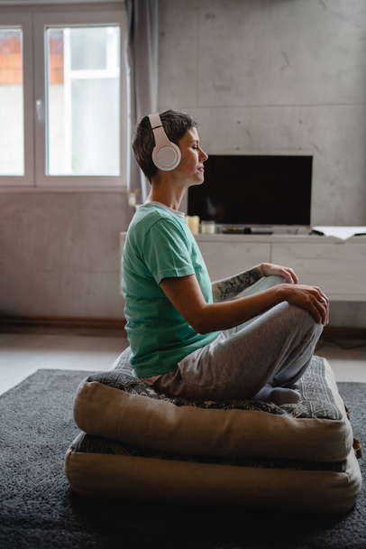 yksi nainen vanhempi valkoihoinen nainen käyttää kuulokkeita verkossa opastettu meditaatio harjoitellaan mindfulness jooga silmät kiinni lattialla kotona oikeita ihmisiä itsehoidon käsite kopioi tilaa - Valokuva, kuva