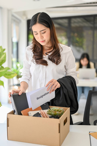 Sad and unhappy young Asian female office worker with a resignation letter in her hand packing her stuff into a cardboard box in the office, quitting her job or getting fired from her boss. - Foto, Imagem