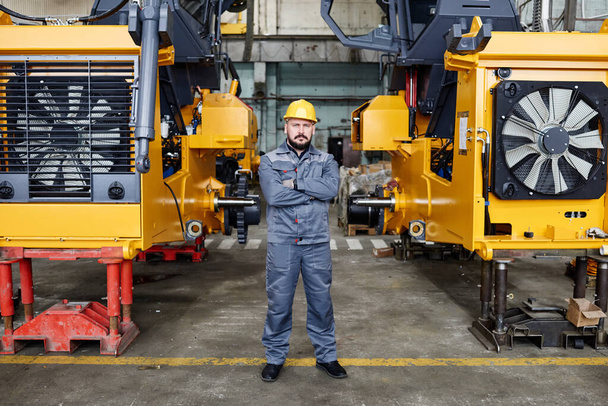 Mature workman in yellow safety helmet and grey boilersuit keeping his arms crossed by chest while standing in warehouse - Fotografie, Obrázek