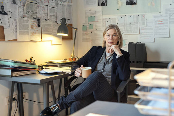 Serious mature female detective with cup of coffee sitting by workplace against walls with criminal profiles and other working documents - Photo, image