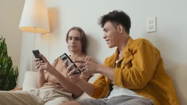 Two buddies in their early 20s scrolling on their smartphones and chatting sitting on couch at home hanging out together - Materiał filmowy, wideo