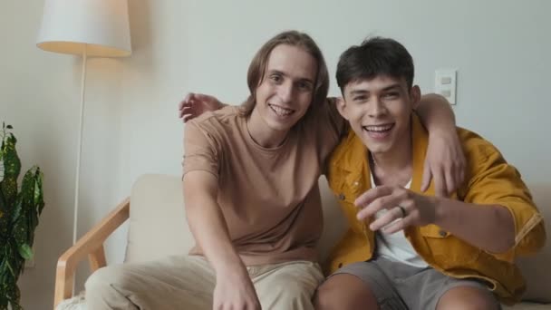 Slowmo portrait of two buddies in their early 20s sitting together on couch indoors pointing and smiling at camera - Materiał filmowy, wideo