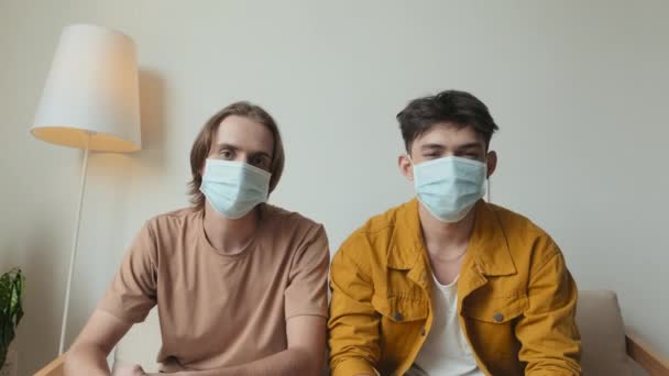 Dolly out portrait of two mates wearing face masks posing for camera sitting on couch indoors - Кадры, видео
