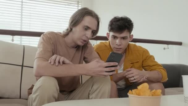 Two mates in their early 20s eating junk food and discussing something on smartphone while hanging out together at home - Materiał filmowy, wideo