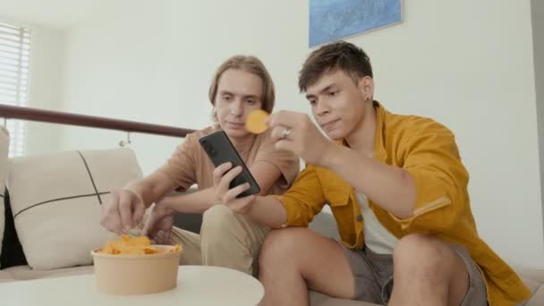 Двоє друзів їдять фішки і обговорюють щось на смартфоні, сидячи на дивані вдома
 - Кадри, відео