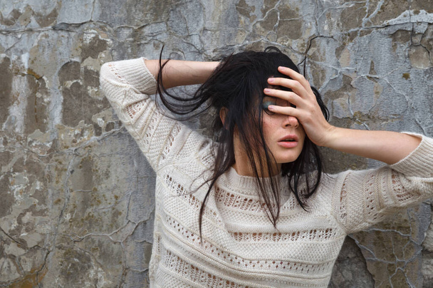 An emotional young woman in a white wool sweater poses against a concrete wall - Fotografie, Obrázek