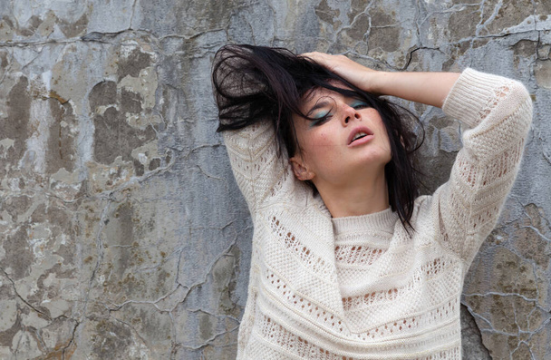 An emotional young woman in a white wool sweater poses against a concrete wall - Foto, Imagem