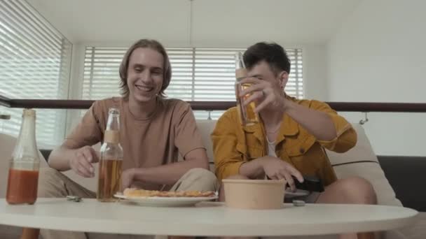 Two buddies in their early 20s drinking beer and watching TV sitting on couch in bright apartment - Materiał filmowy, wideo