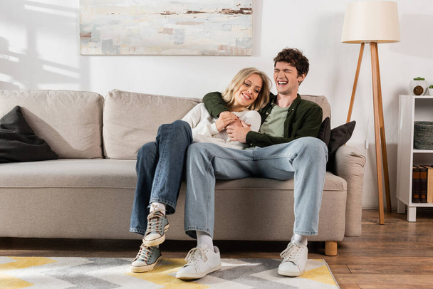 full length of cheerful and curly man hugging happy blonde girlfriend in living room  - Φωτογραφία, εικόνα