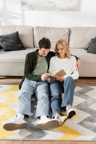 full length of cheerful couple smiling while reading book together in living room  - Foto, Imagen