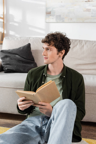 intelligent man with curly hair holding book while looking away in living room  - Foto, afbeelding