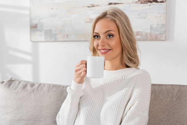 dreamy young woman in white sweater holding cup of coffee and smiling at home  - Valokuva, kuva