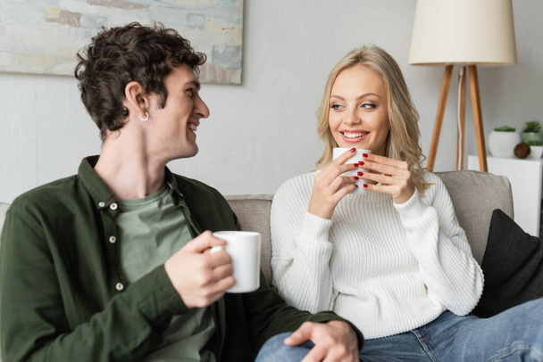 happy young man and blonde woman in sweater holding cups with coffee in living room  - Fotó, kép