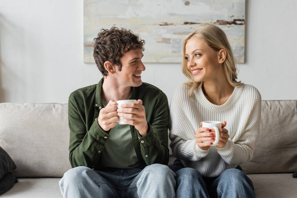 cheerful young man and blonde woman holding cups with coffee while looking at each other in living room  - Foto, Imagem