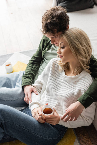 high angle view of young man with curly hair hugging blonde woman with cup of tea in living room  - Foto, imagen
