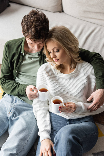 high angle view of young man with curly hair hugging blonde woman while holding cups of tea in living room  - Fotoğraf, Görsel