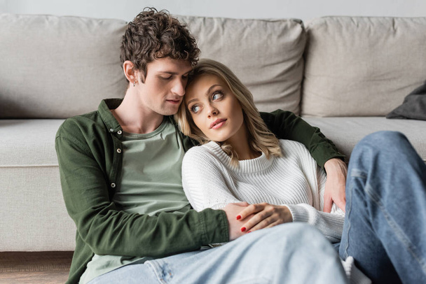 young man with curly hair hugging blonde woman near sofa in living room  - Fotó, kép