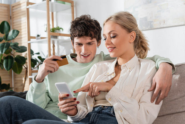 cheerful woman pointing at smartphone while doing online shopping near boyfriend holding credit card  - Foto, imagen