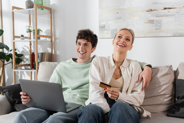 cheerful woman holding credit card near boyfriend with laptop doing online shopping  - Fotoğraf, Görsel