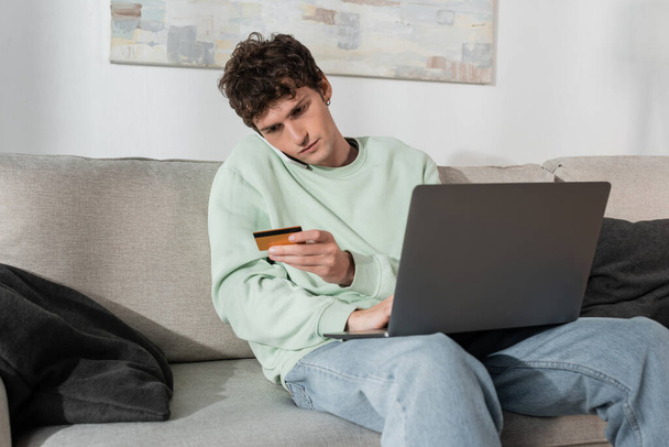 curly young man holding credit card while talking on smartphone near laptop  - 写真・画像